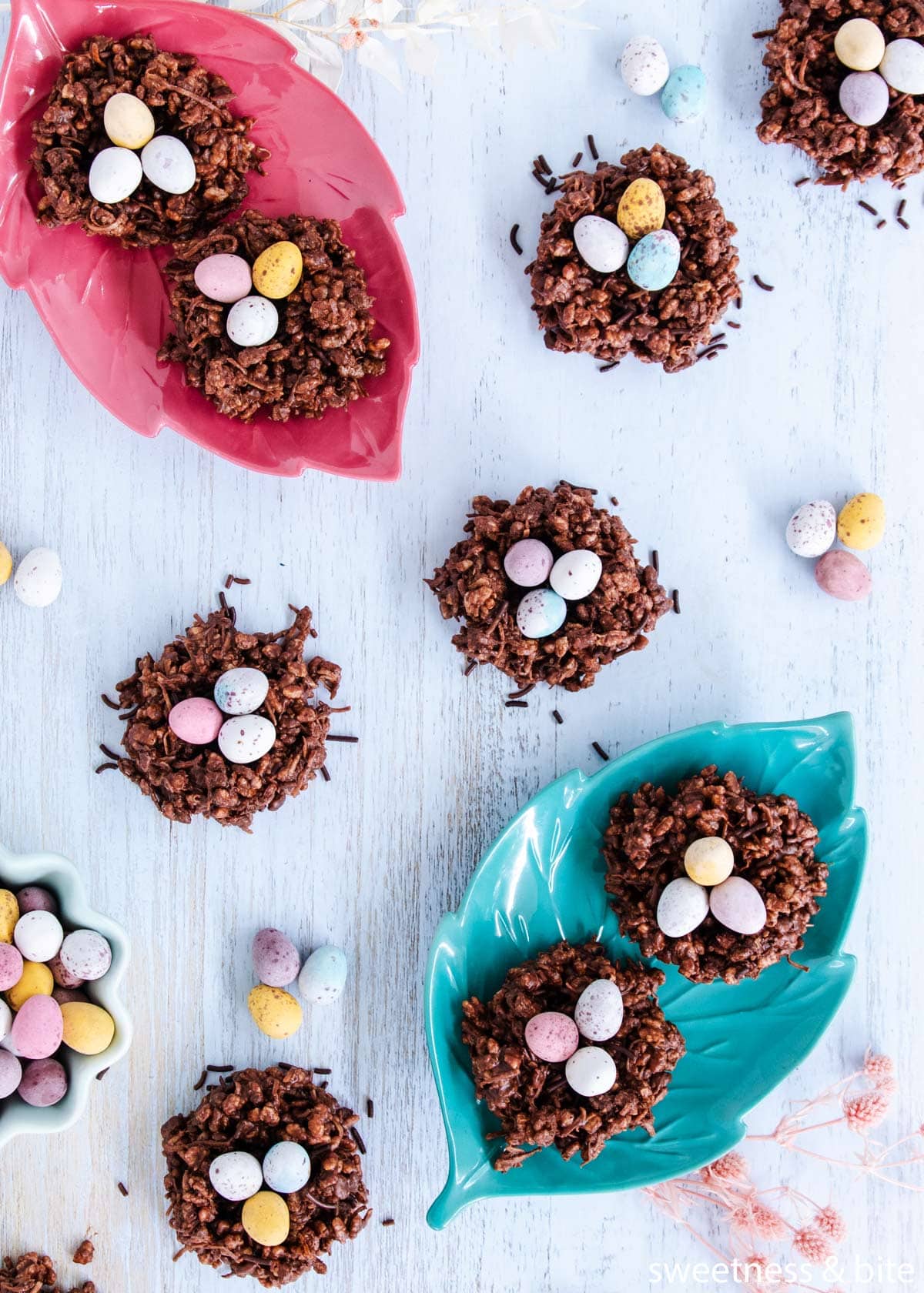 Overhead shot of nests, some on leaf-shaped plates, on a blue woodgrain background, surrounded by pastel eggs.