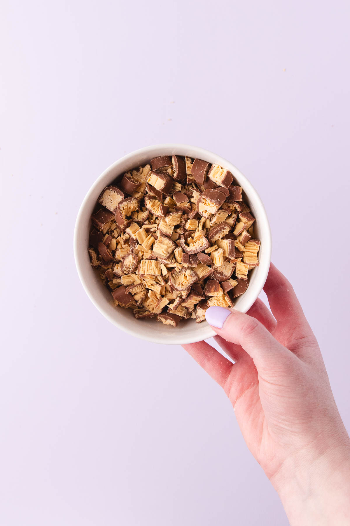 A hand holding a small white bowl of chopped Caramilk Twirls.