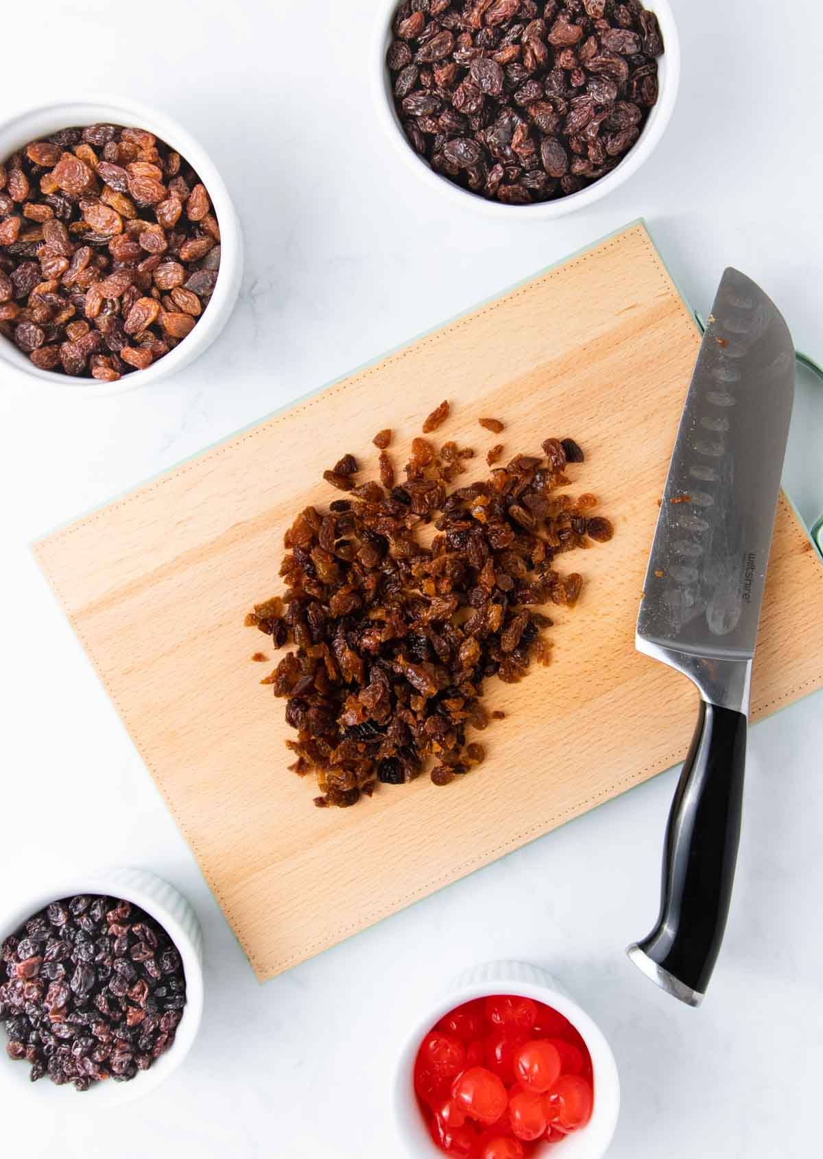 Chopped raisins on a wooden chopping board with a large kitchen knife and other dried fruit in small white bowls.