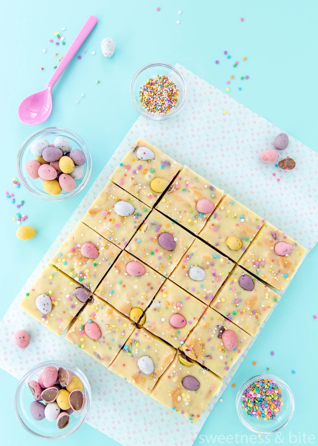 Mini egg fudge slice cut into 16 squares, with sprinkles and mini eggs on a pale blue background.