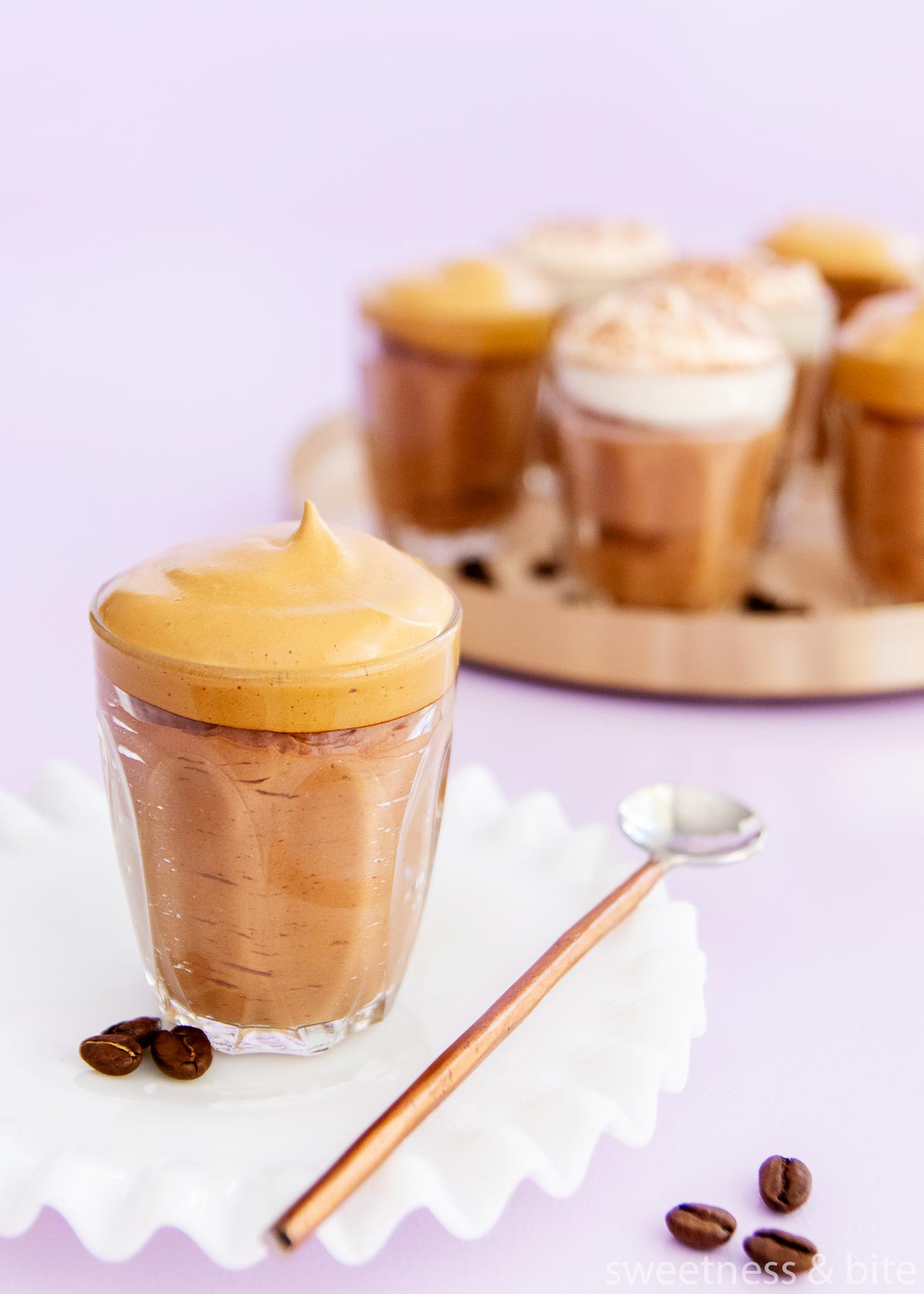 A small glass of mocha mousse with whipped coffee topping on a white milk glass plate with a ruffled edge, and a copped handled spoon.