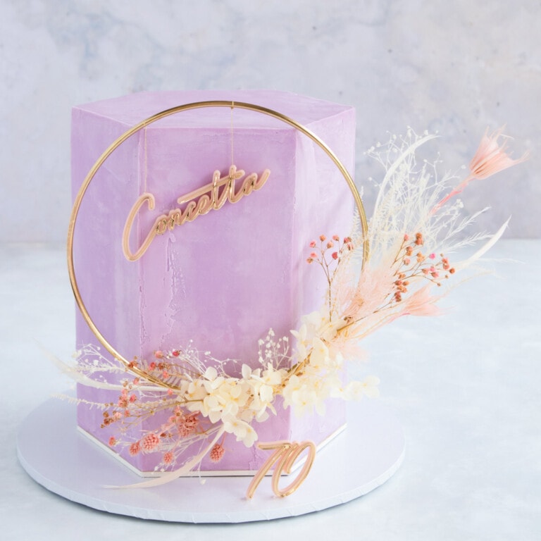 A tall hexagon shaped cake covered in lilac ganache, with a pink and white floral cake topper.
