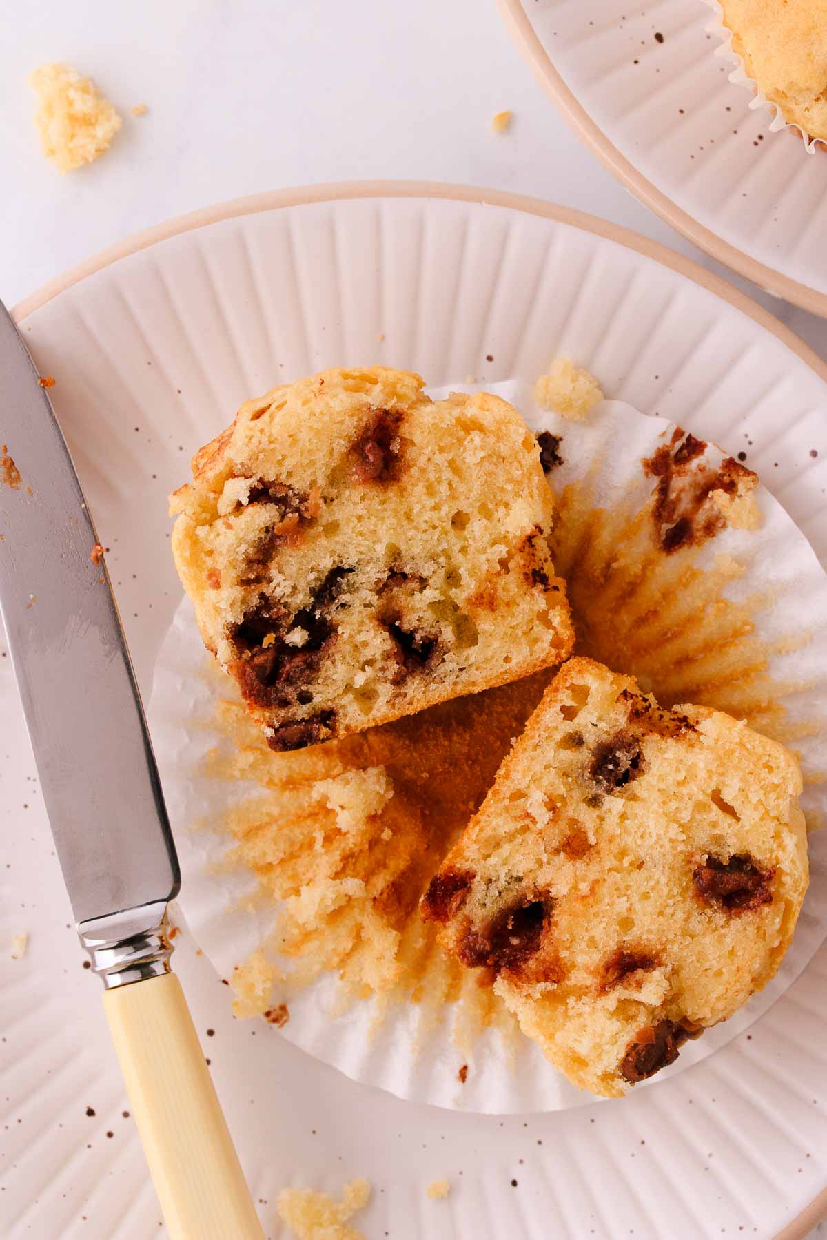 A halved chocolate chip muffin showing the fluffy texture, on a white ridged plate, with a bone-handled knife.