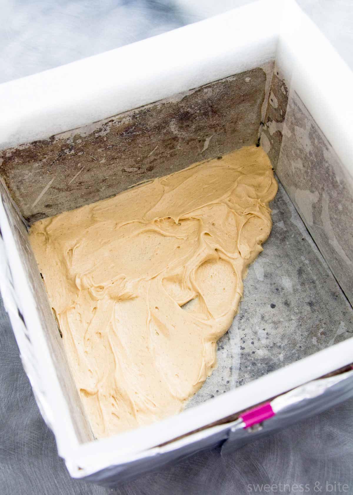 A square cake pan lined with baking paper, with cake batter spread over the bottom.