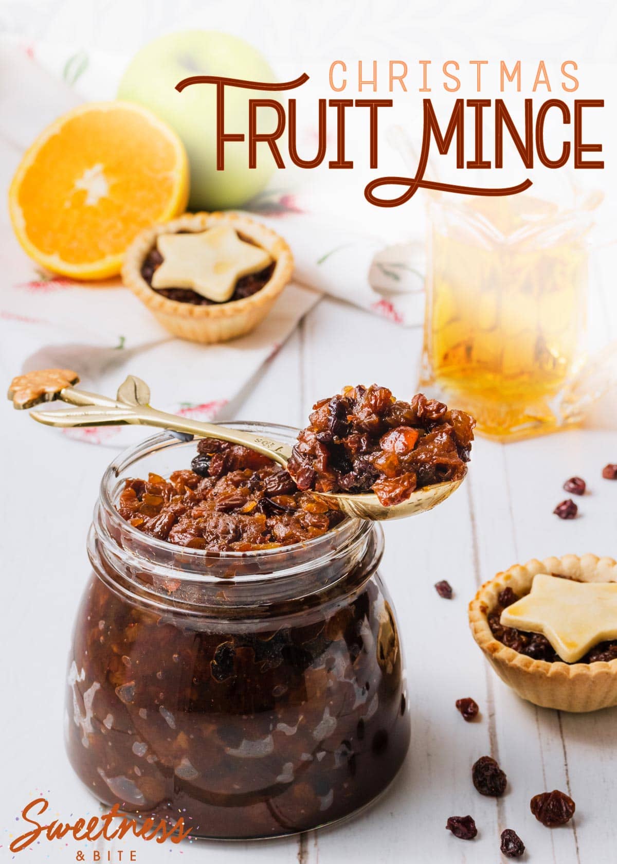 A large glass jar of fruit mince, on a white wooden background, with fruit mince pies, an apple, an orange and a glass jug of brandy, text overlay reads 