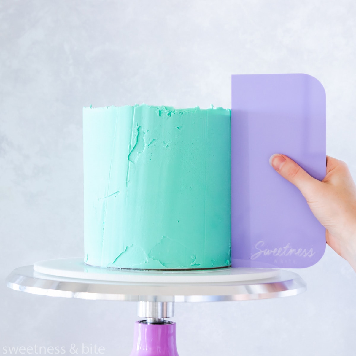 A cake being ganached with teal ganache using a purple scraper