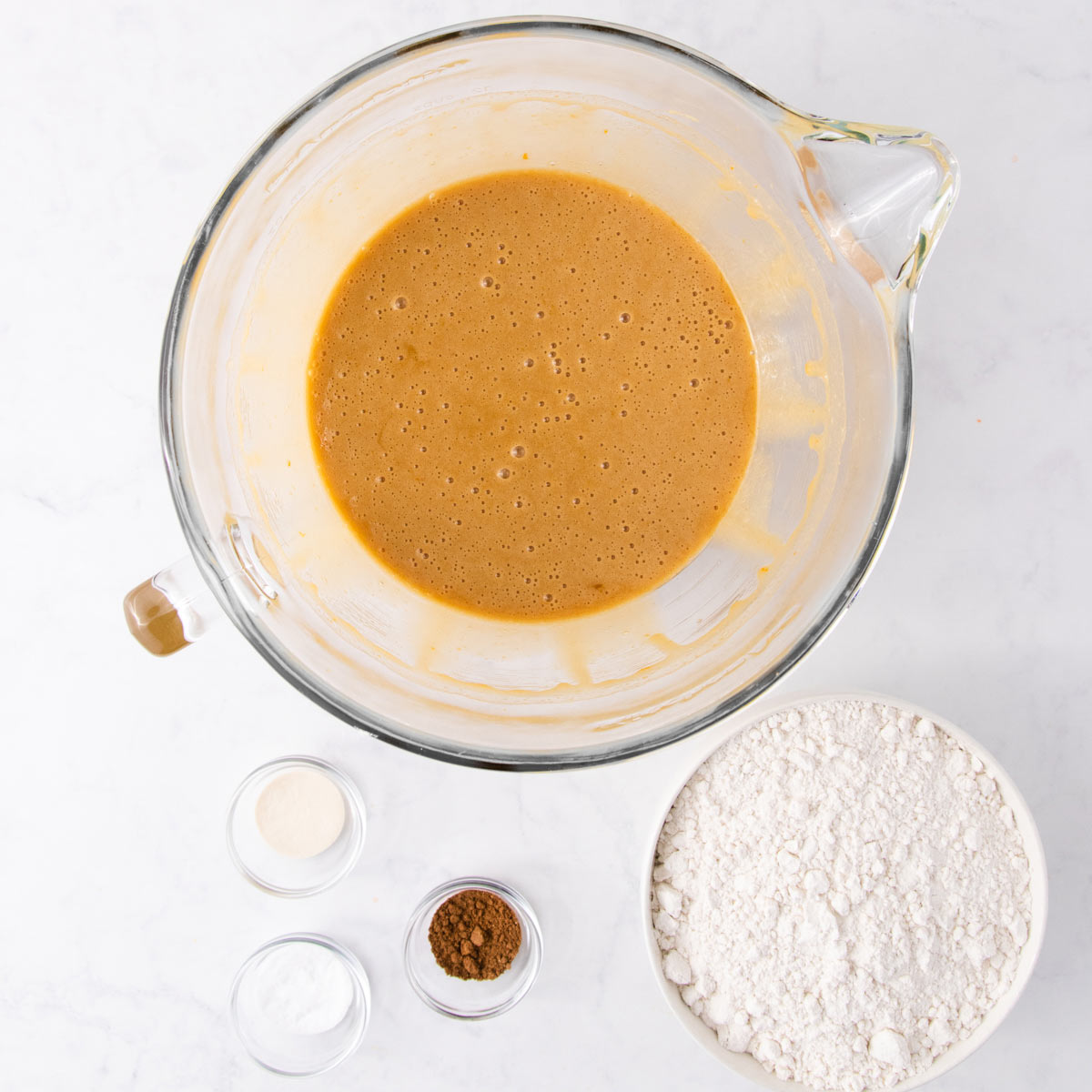 The beaten wet ingredients in a glass mixer bowl, with the dry ingredients in small bowls, on a grey background.