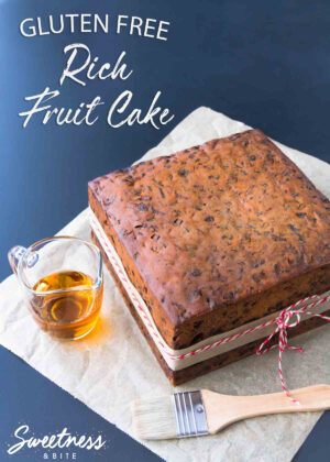 A square gluten free rich fruit cake on a piece of parchment paper, with a small glass jug of brandy and a pastry brush