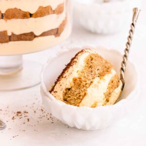 Gluten free tiramisu in a footed glass trifle bowl, on a grey/blue background, with small white bowls and a serving spoon.