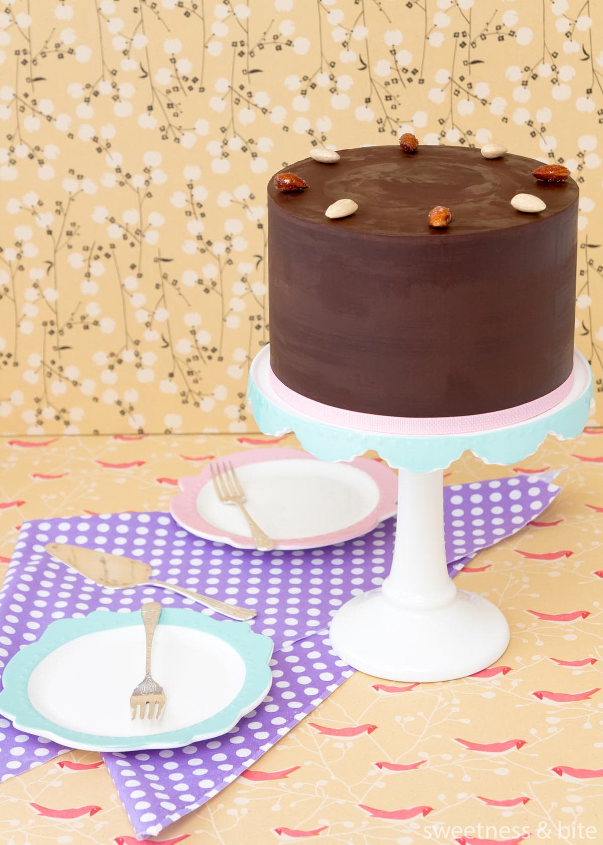 A dark chocolate ganache-covered cake decorated with blanched and honey roasted almonds, on a white and blue cake stand,