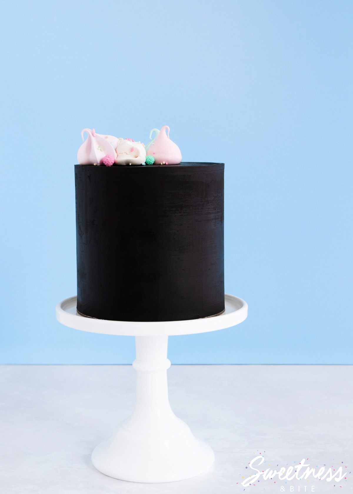A cake covered in black chocolate ganache, on a white cake stand, topped with pastel meringues.