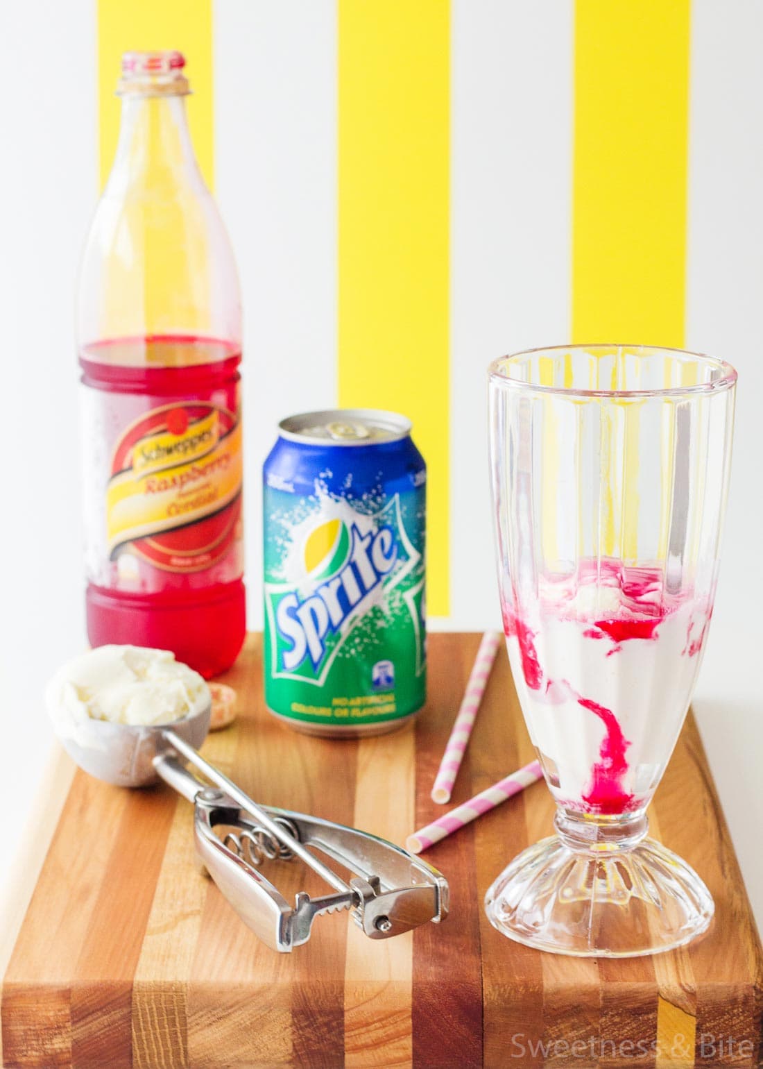 A tall glass with a scoop of ice cream and some raspberry cordial, with a bottle of cordial and a cake of lemonade in the background.