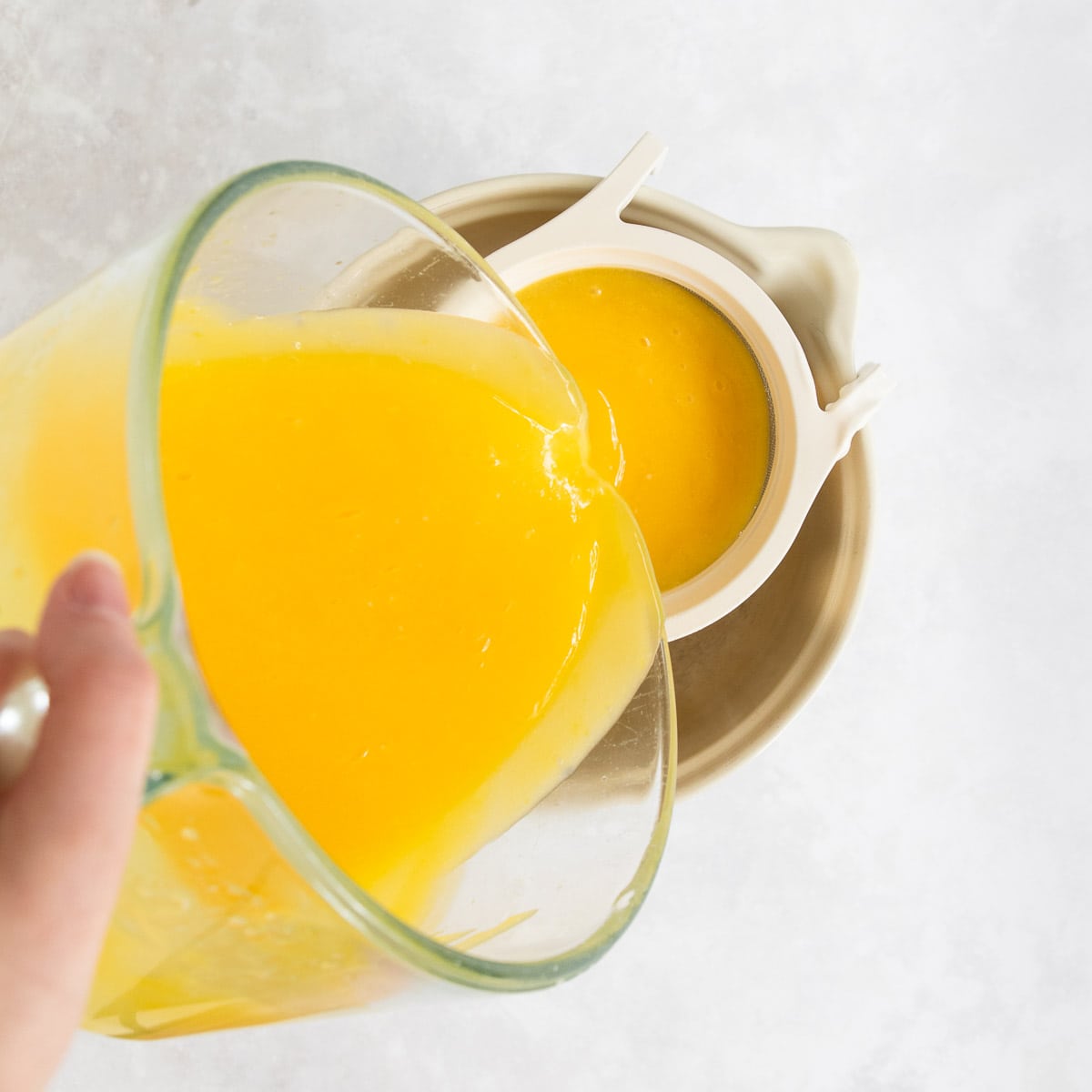 The lemon curd being poured through a fine mesh sieve into a large ceramic jug.