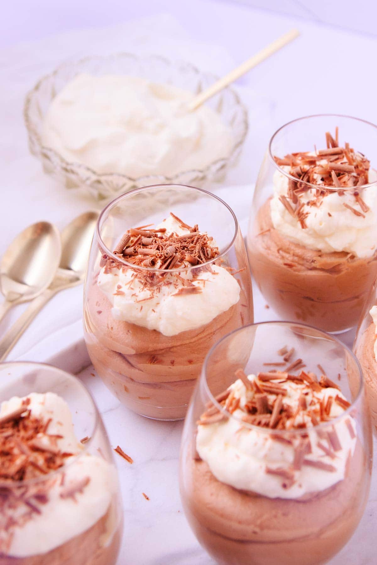 Four glasses of mousse, with a small glass bowl of whipped cream, and gold teaspoons in the background.