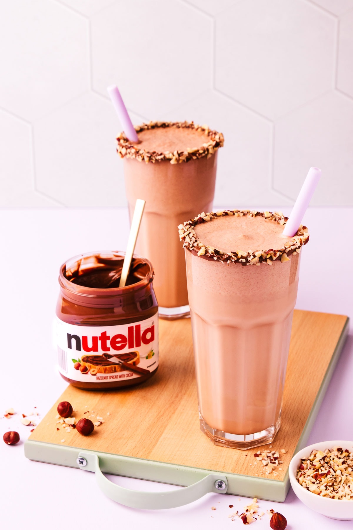 Two Nutella milkshakes on a small wooden chopping board, with an open jar of Nutella and scattered chopped and whole hazelnuts.