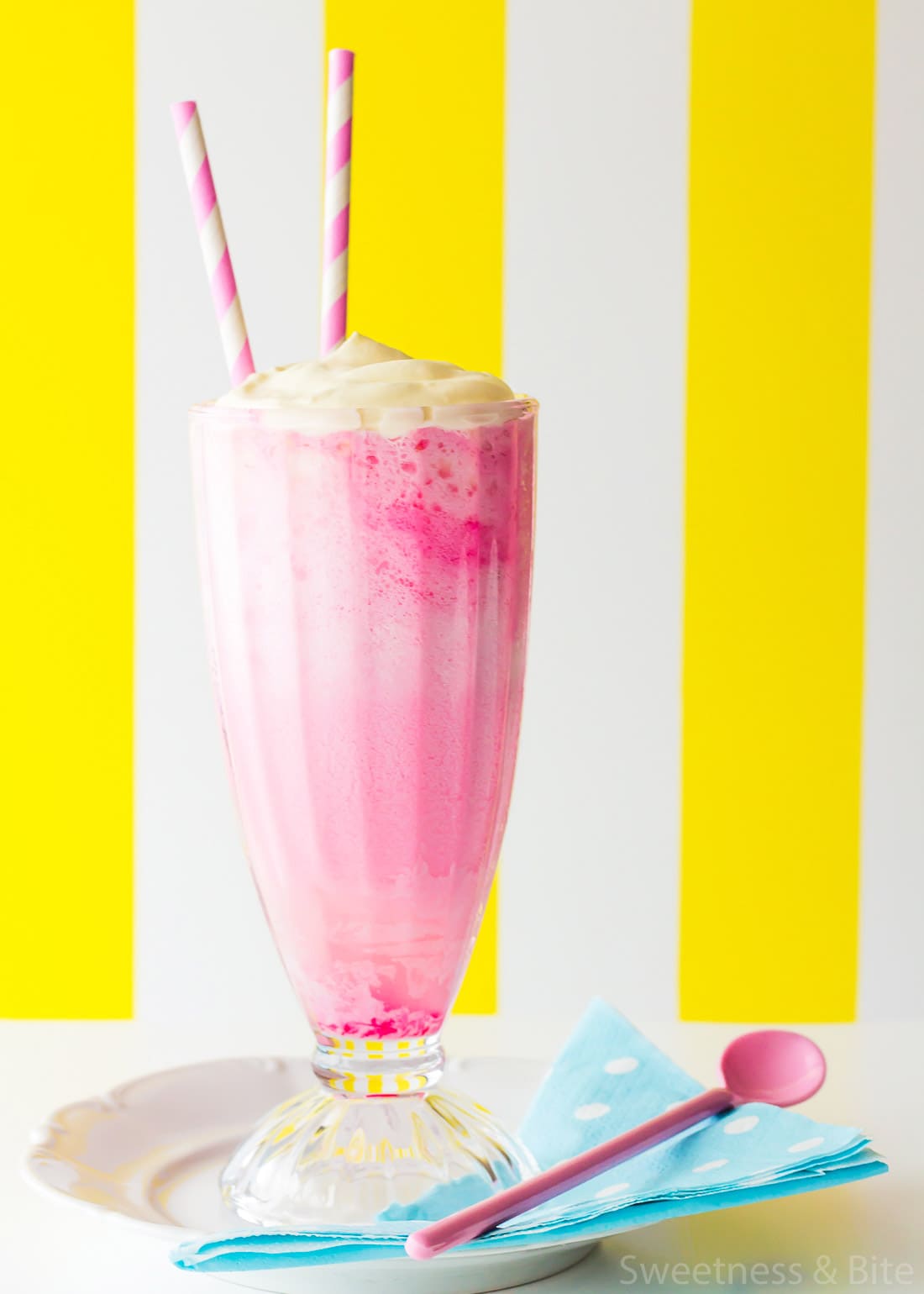 Close up of the pink panther drink in a tall glass, sitting on a white plate with a blue spotted paper napkin and a pink spoon.