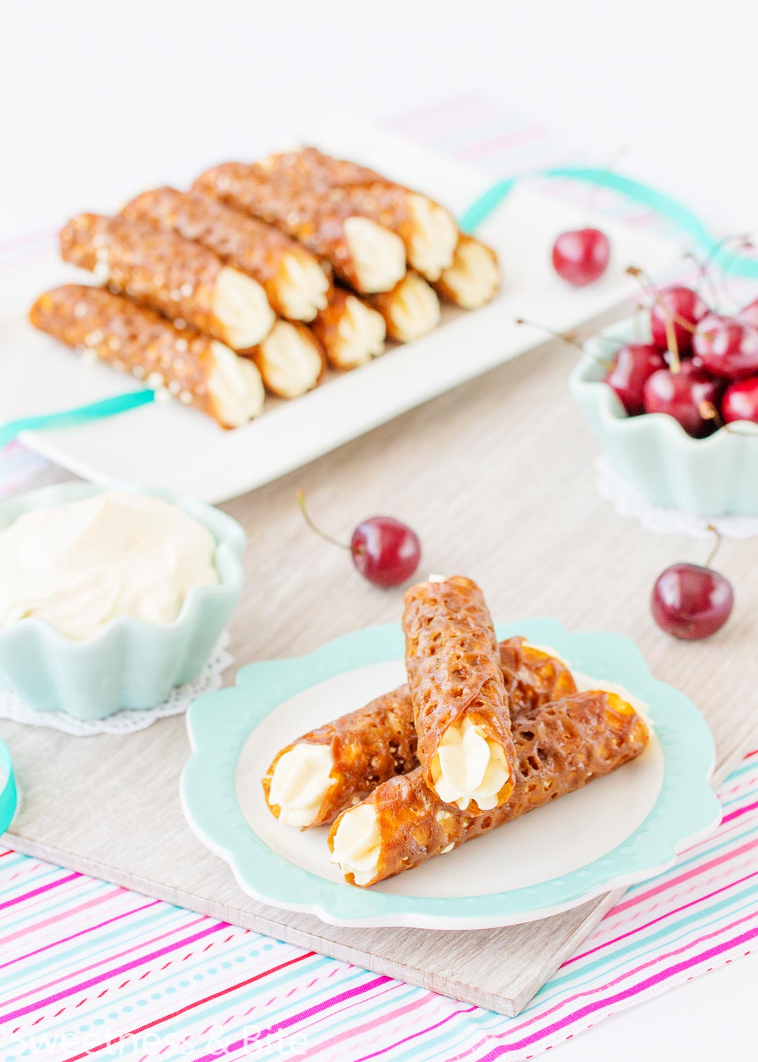 Three gluten free brandy snaps stacked on a plate in the foreground, with a bowl of whipped cream and fresh cherries in the background.