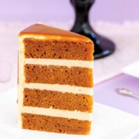 A slice of rich caramel mud cake with dripping caramel sauce on a white plate with a purple background.