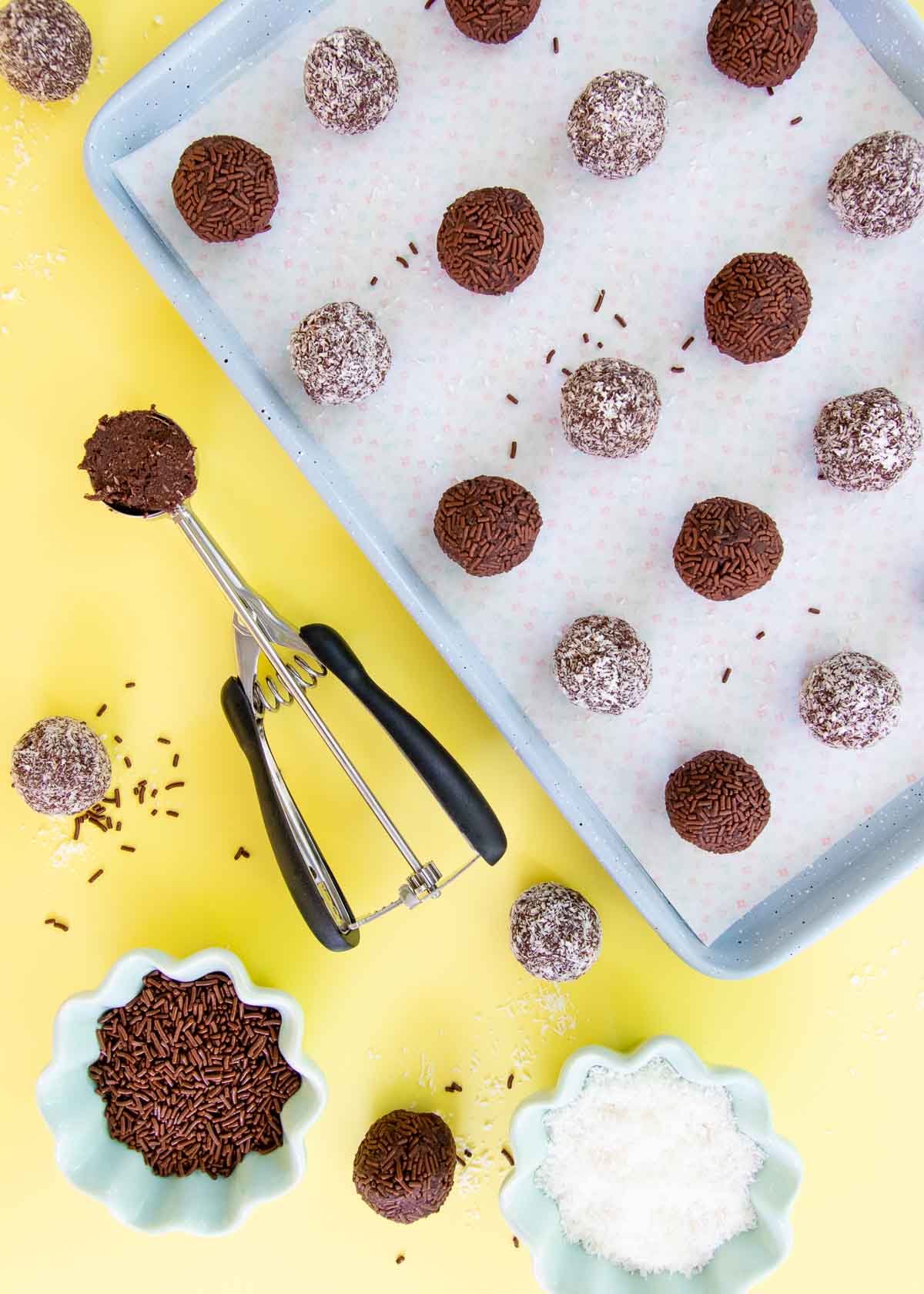 A tray of truffles, with a small cookie scoop, and small bowls of coconut and chocolate sprinkles, on a yellow background.