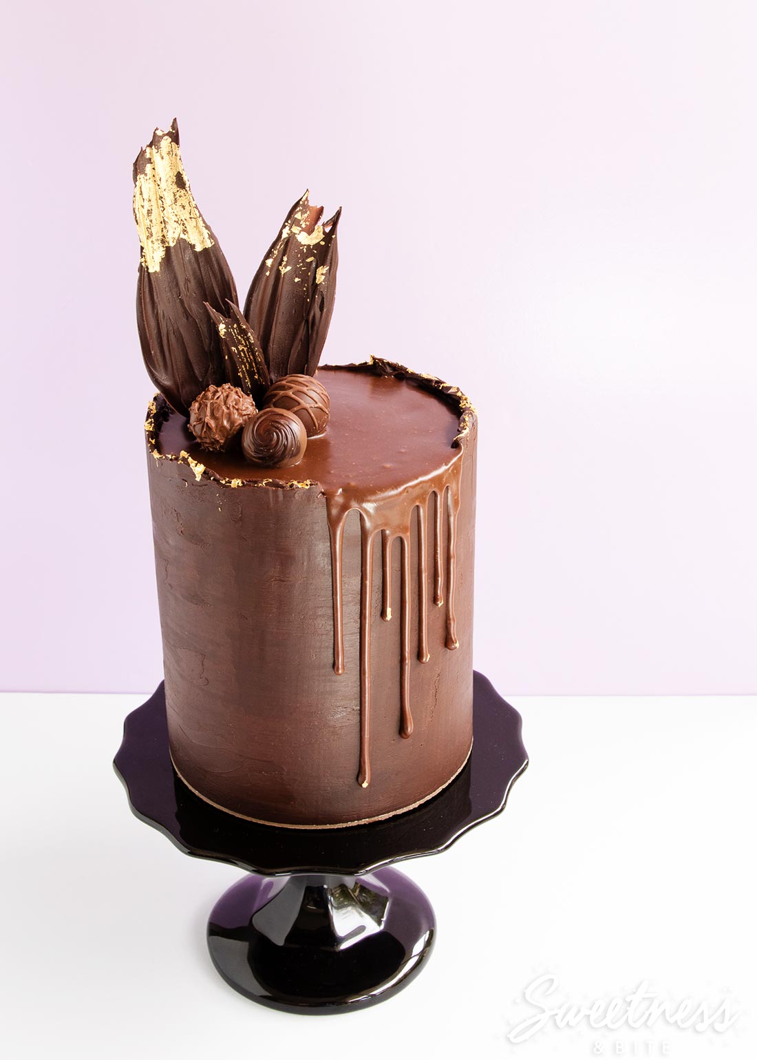 A ganache covered cake with a rough top edge and gold leaf, topped with a ganache drip and chocolate shards.