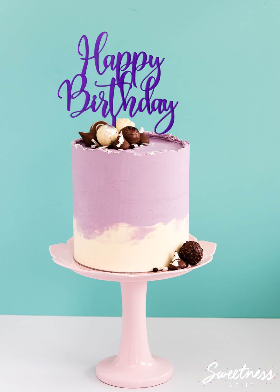 A cake covered in purple and white swirled ganache with a rough top edge, decorated with chocolates and a happy birthday acrylic topper.