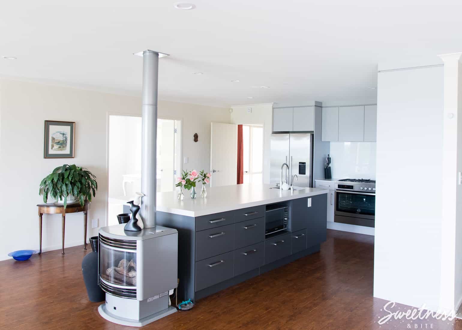 A kitchen with light and dark grey cabinetry and a very wide and long island bench, with a gas fireplace at one end.