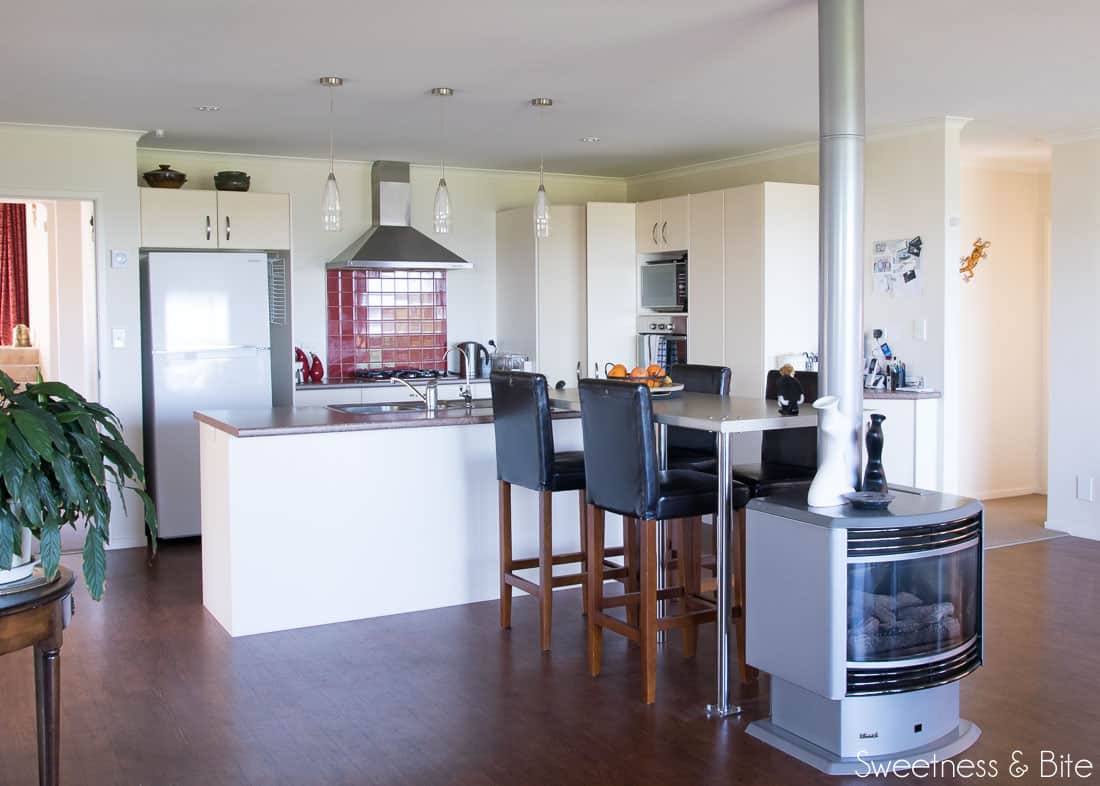 An ugly old kitchen with a red tile splashback, a very small bench, a long, too high breakfast bar, and a gas fireplace at the end. 