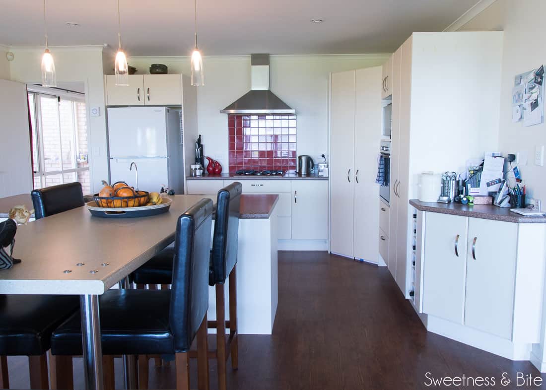 An ugly old kitchen with a red tile splashback, a very small bench, a long, too high breakfast bar, and a gas fireplace at the end (view from the opposite side.)