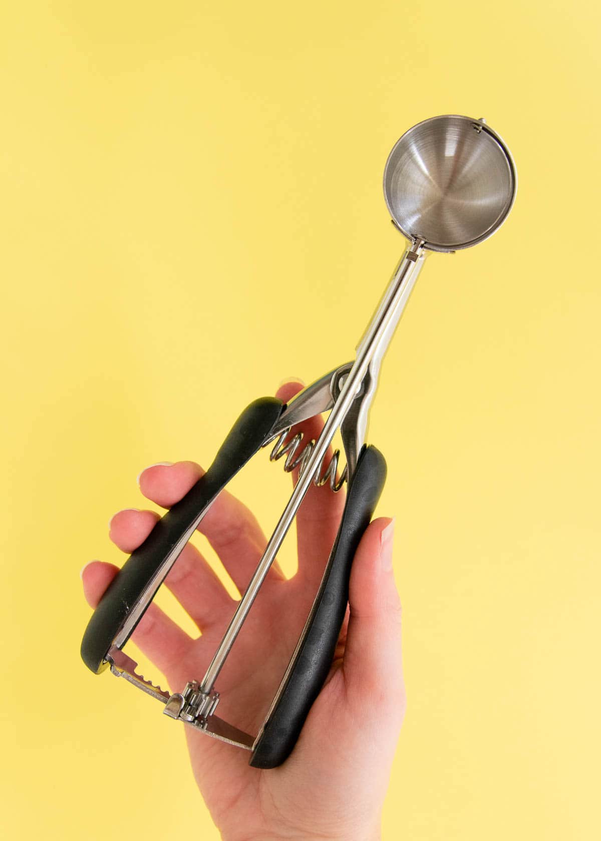 A hand holding up a 1 tablespoon capacity cookie scoop, with a yellow background.