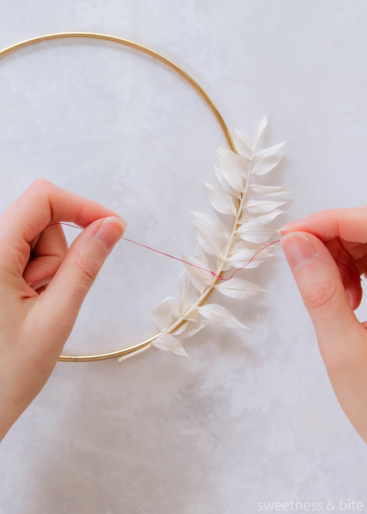 A stem of white ruscus leaved being tied to the hoop with pink thread.