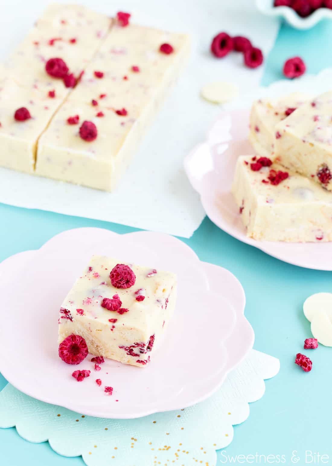 One piece of white choc raspberry slice on a pink flower shaped plate in the foreground. Other pieces of slice on a piece of parchment in the background, on a blue-green tabletop.