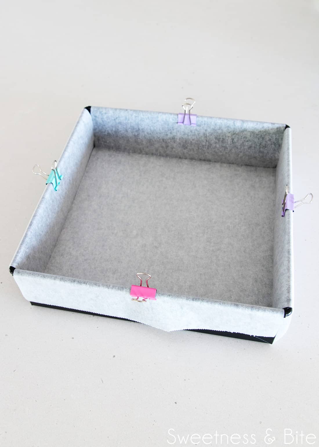 A dark metal square cake pan, with two strips of baking paper criss-crossed to line the pan, with coloured clips on each edge to hold the paper in place.